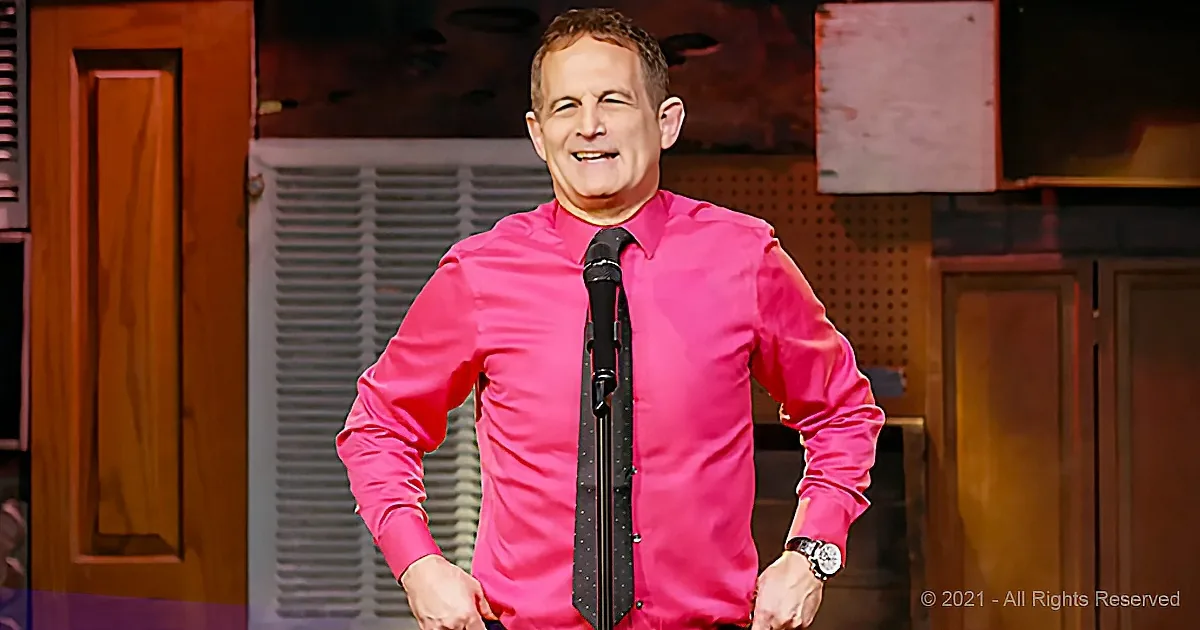 A man in a bright pink shirt and black tie stands on stage with a microphone, smiling.