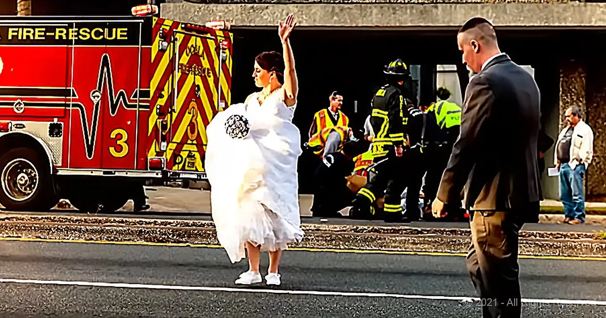A bride in a white dress stands on a street, raising her hand, with a fire truck and emergency personnel in the background.