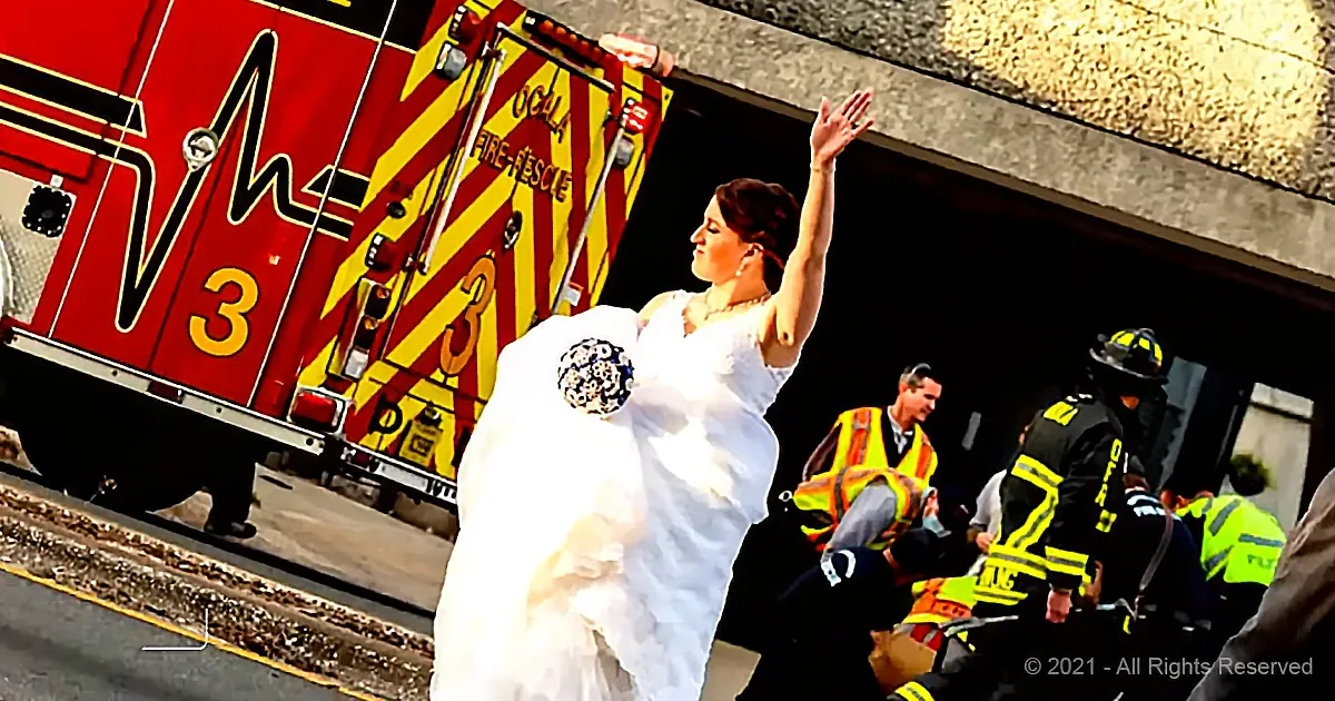 A bride in a white wedding dress raises her hand, standing in front of a fire truck with emergency personnel nearby.
