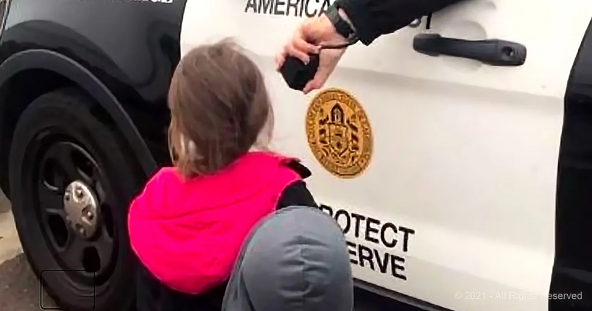 A child in a pink hoodie interacts with a police officer near a vehicle.