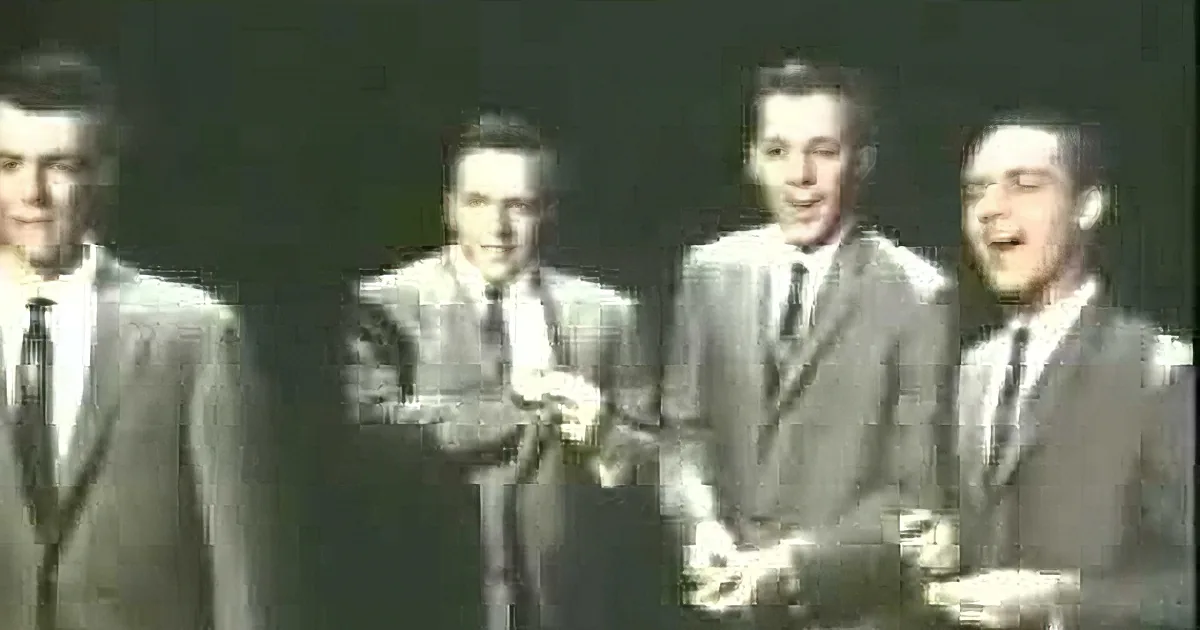 A man in a suit and tie speaks into a microphone, with another person in the background.