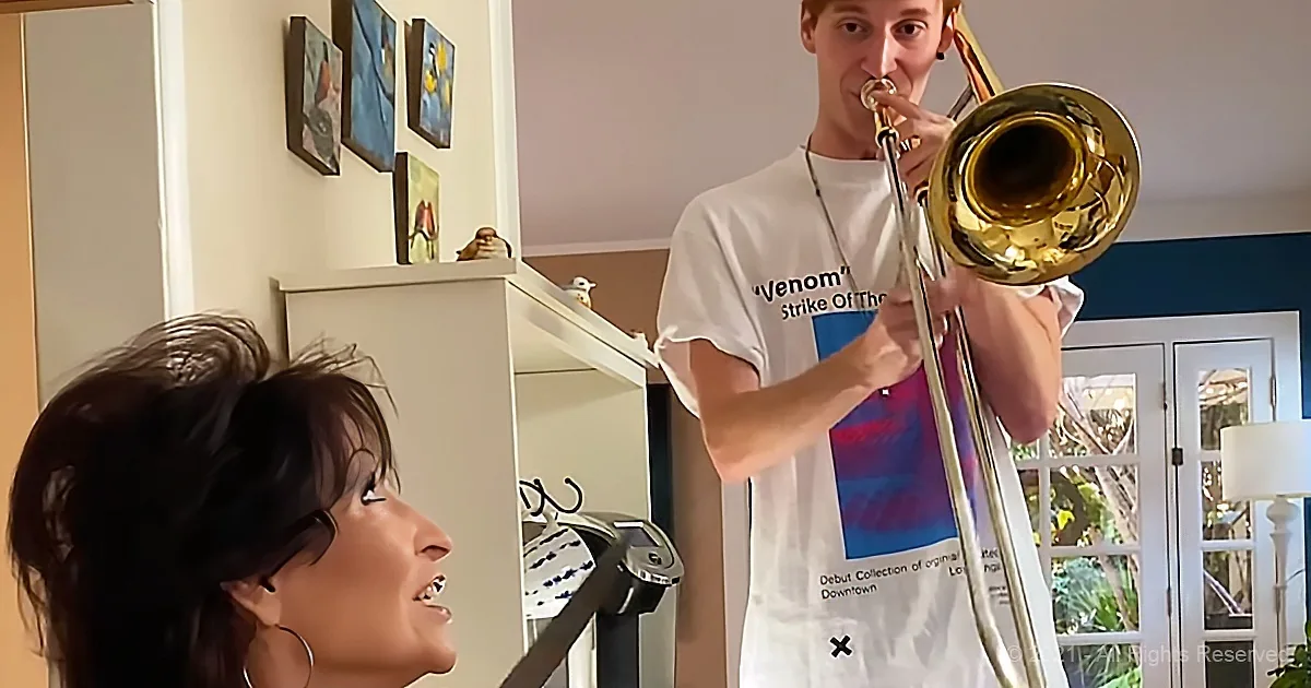 A man plays a trombone while a woman smiles, both in a cozy room with artwork and furniture.