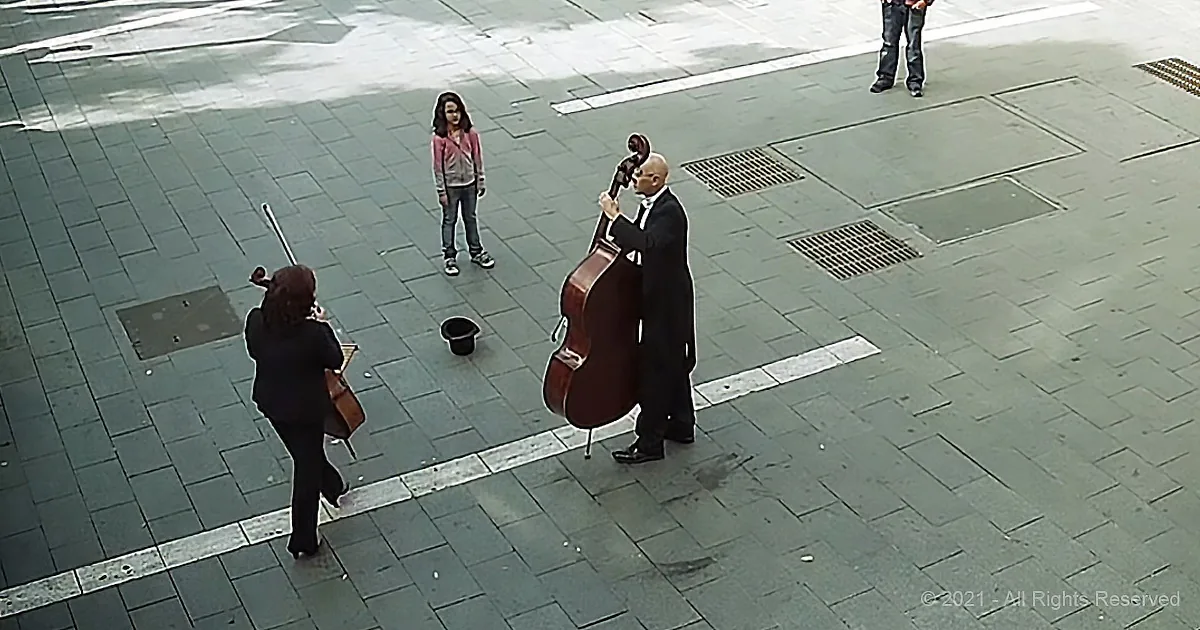 A man in a suit plays a cello in a public square, with a woman and another musician nearby.