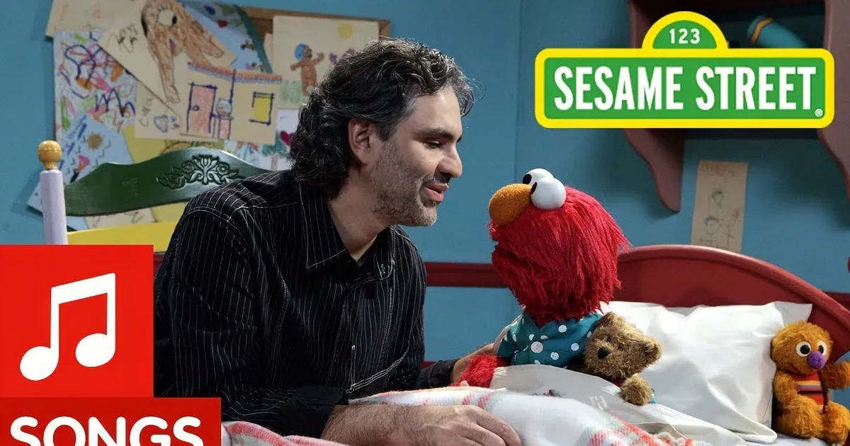 A man with curly hair and a striped shirt is speaking, with a colorful background featuring children's drawings.