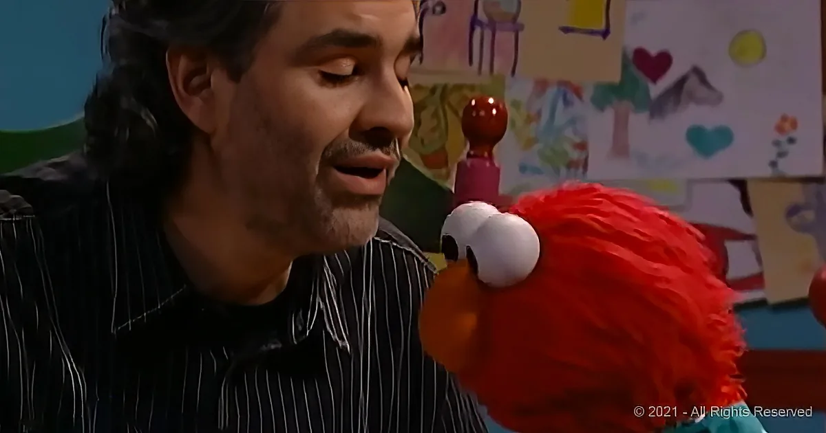 A man in a striped shirt interacts with Elmo, a red puppet, in a colorful setting.