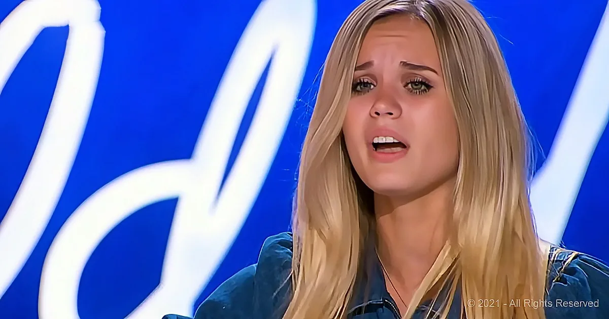 A woman with long blonde hair, wearing a denim jacket, appears to be speaking or reacting, with a blue background...