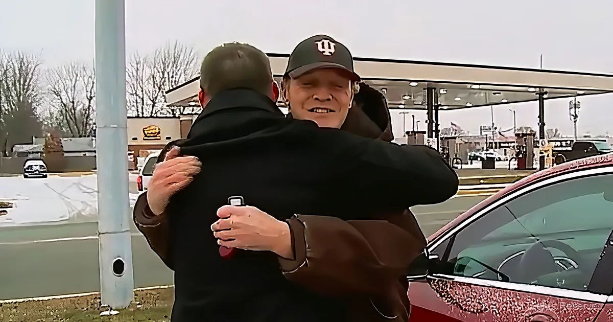 A man in a black coat and cap hugs another man in a parking lot near a gas station.