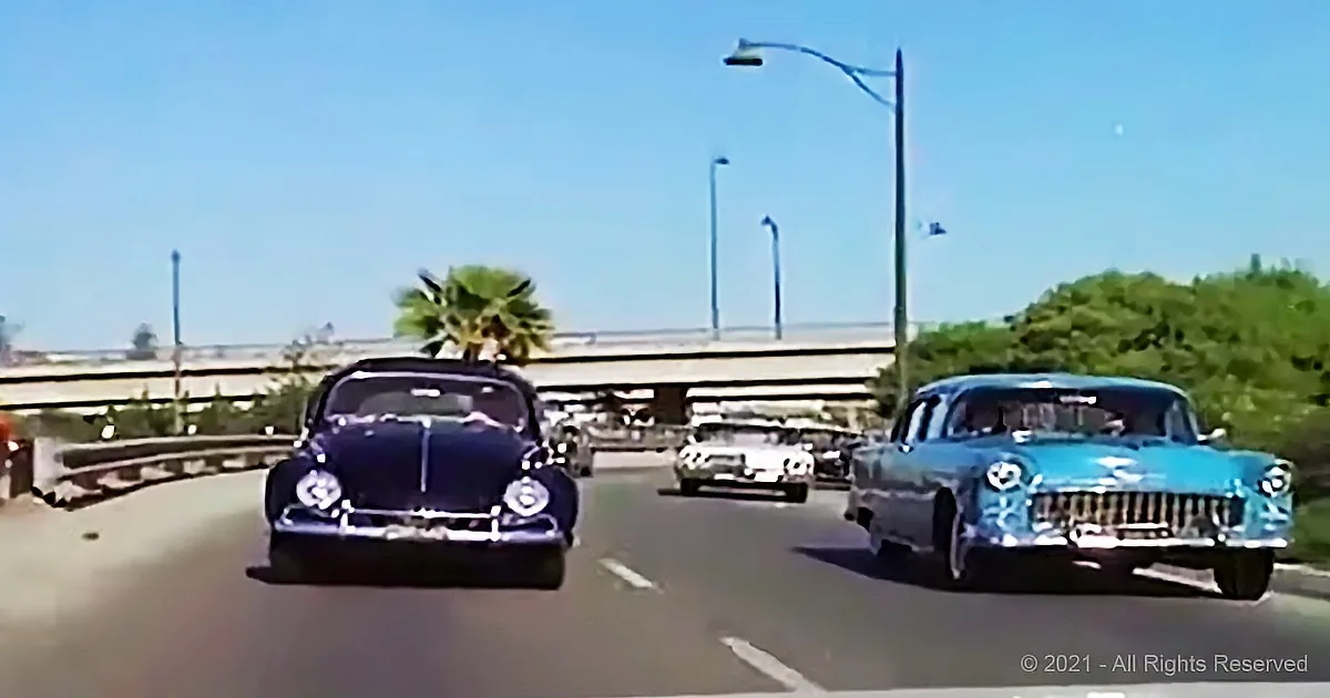 Classic cars drive on a sunny road with palm trees and a bridge in the background.