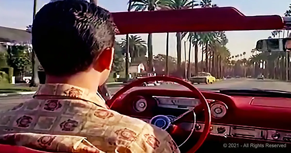 A man in a patterned shirt drives a red convertible car with palm trees and a sunny street in the background.