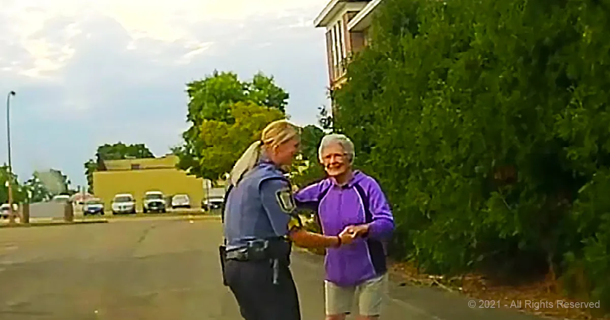 A police officer and a man in a purple shirt are shaking hands on a street with trees and parked cars in the background.