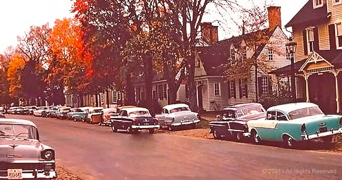 Vintage cars line a street with autumn trees and classic houses in the background.
