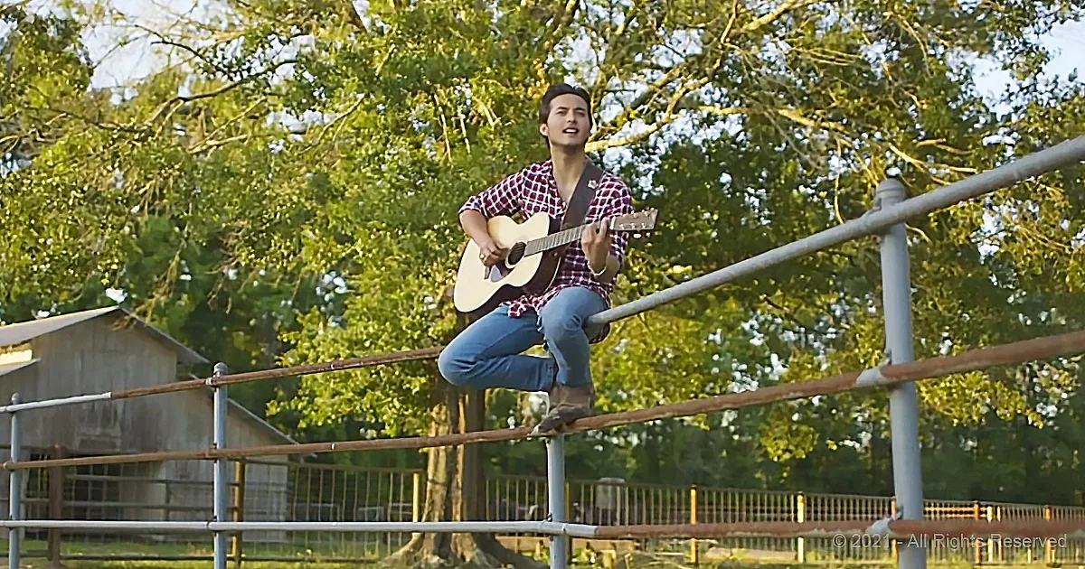 A person in a plaid shirt plays guitar on a fence, surrounded by trees and a barn.