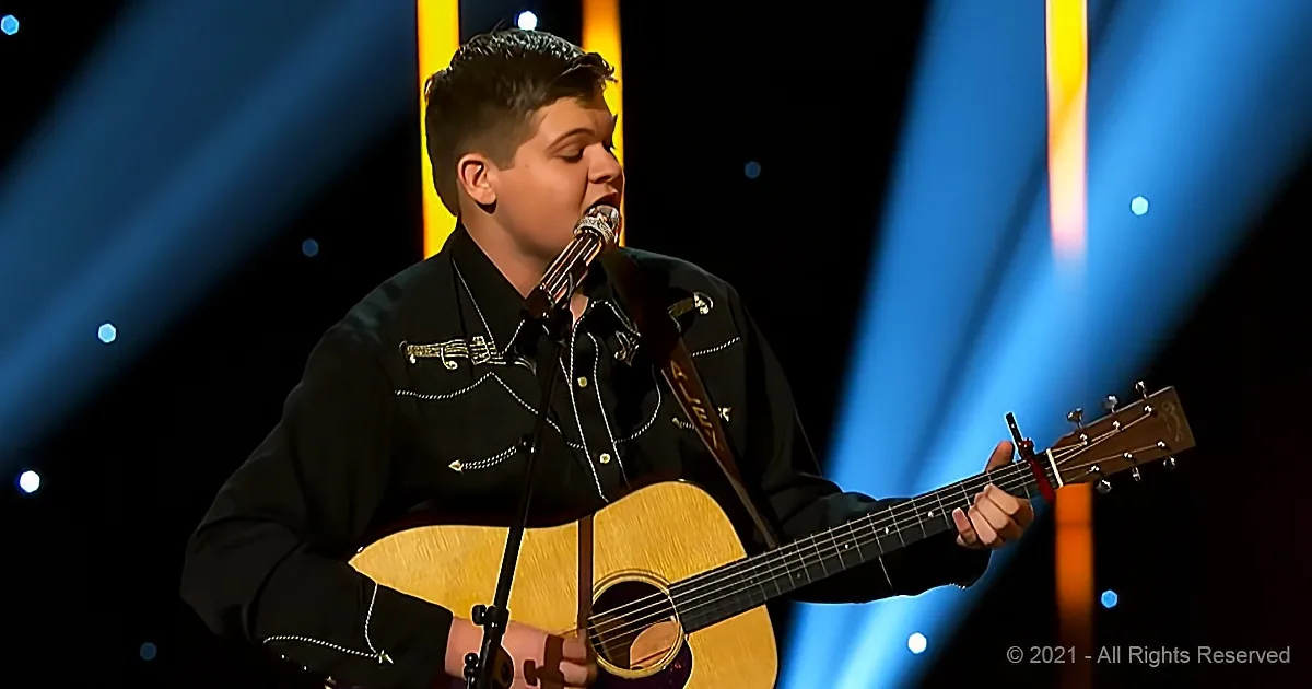 A person in a black outfit plays an acoustic guitar on stage with a microphone.