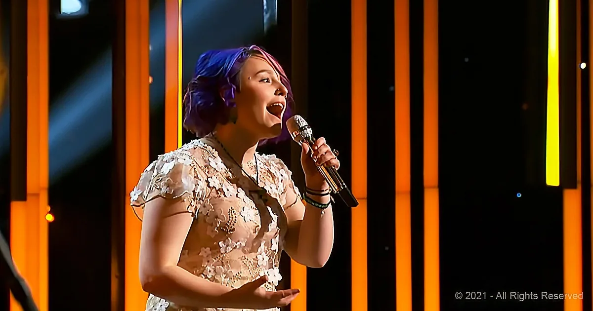 A person with purple hair sings into a microphone, wearing a floral dress, against an orange-lit background.