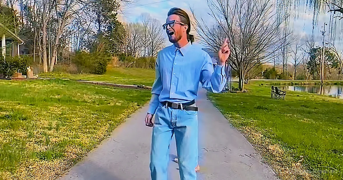 A man in a light blue shirt and jeans stands on a path, gesturing with his right hand.