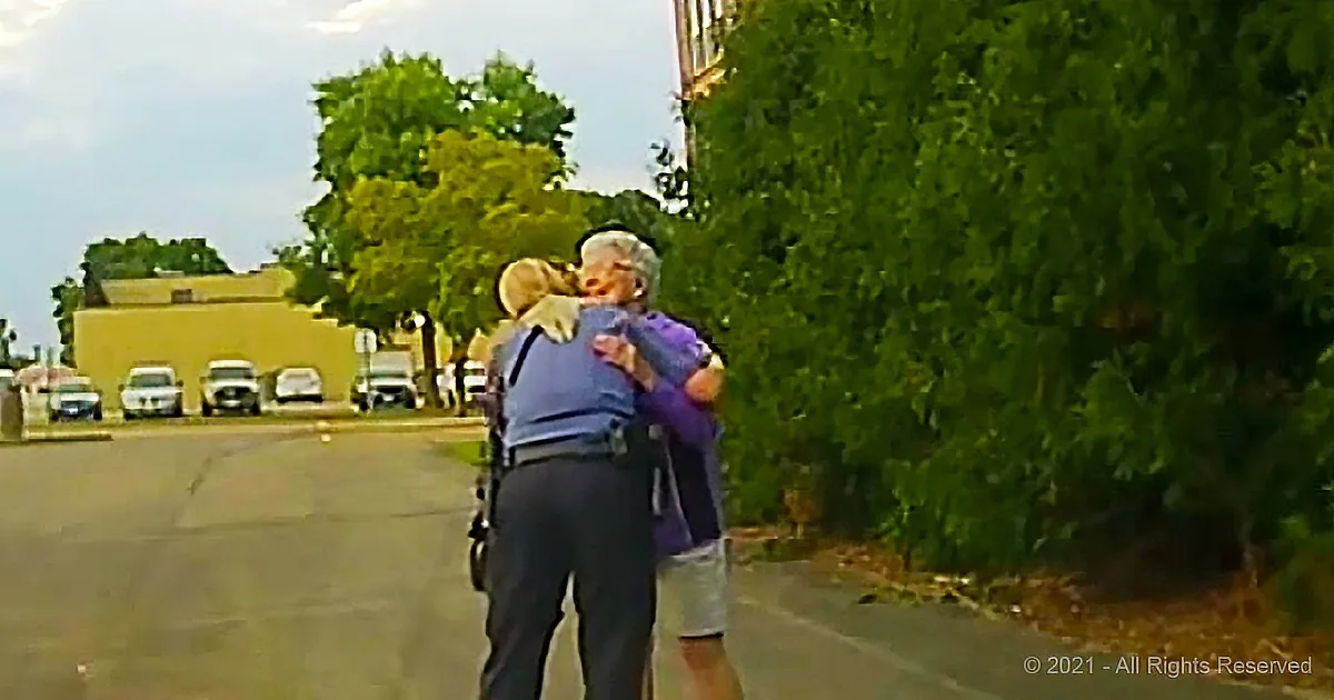 A police officer hugs an elderly woman in a parking lot near trees and a building.