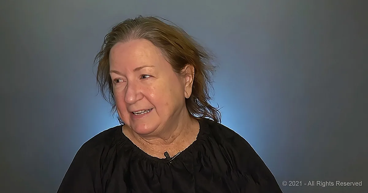 An older woman with shoulder-length hair, wearing a black top, smiles while speaking into a microphone.