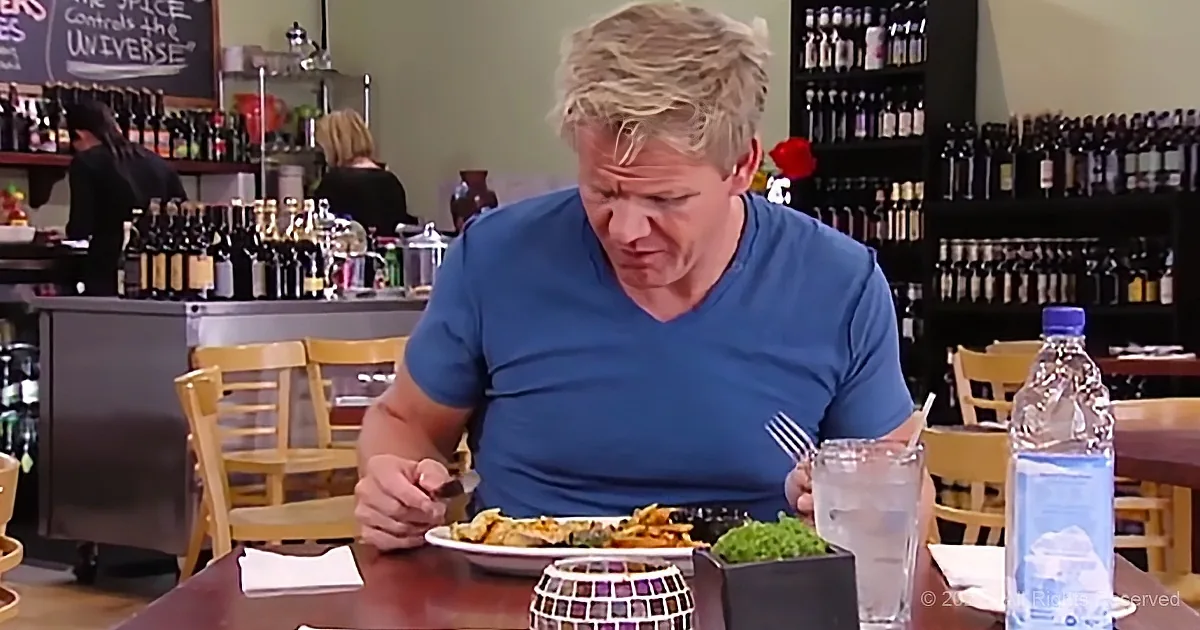 A man in a blue shirt sits at a table with a plate of food, surrounded by bottles and bar elements.