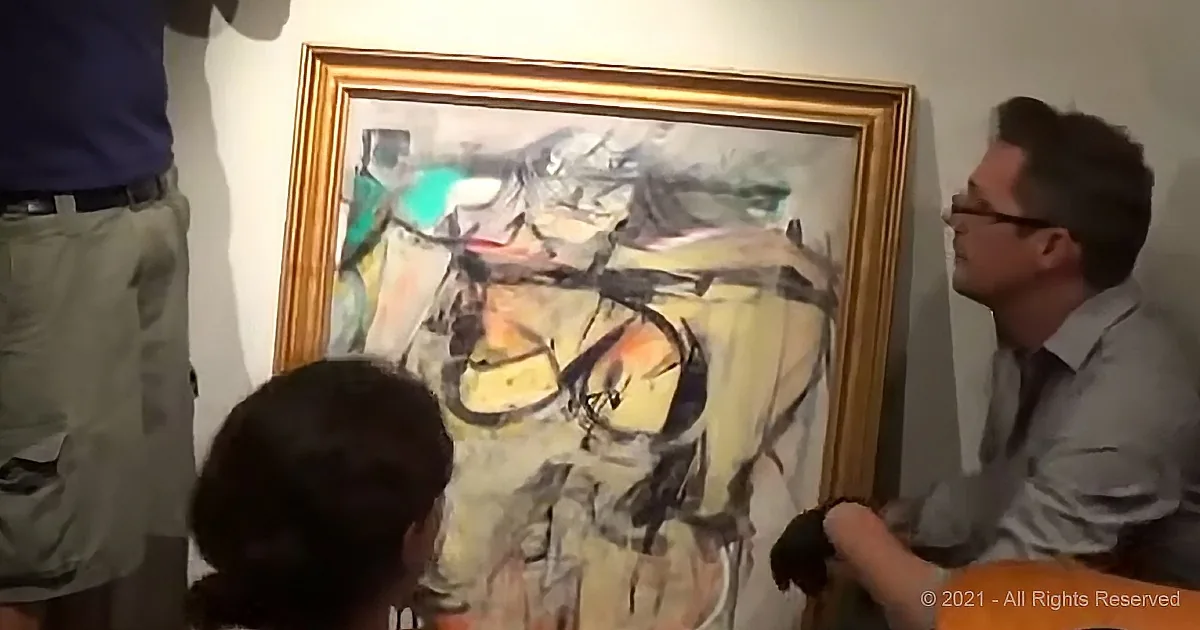 A man in glasses examines an abstract painting with a group of people around it.