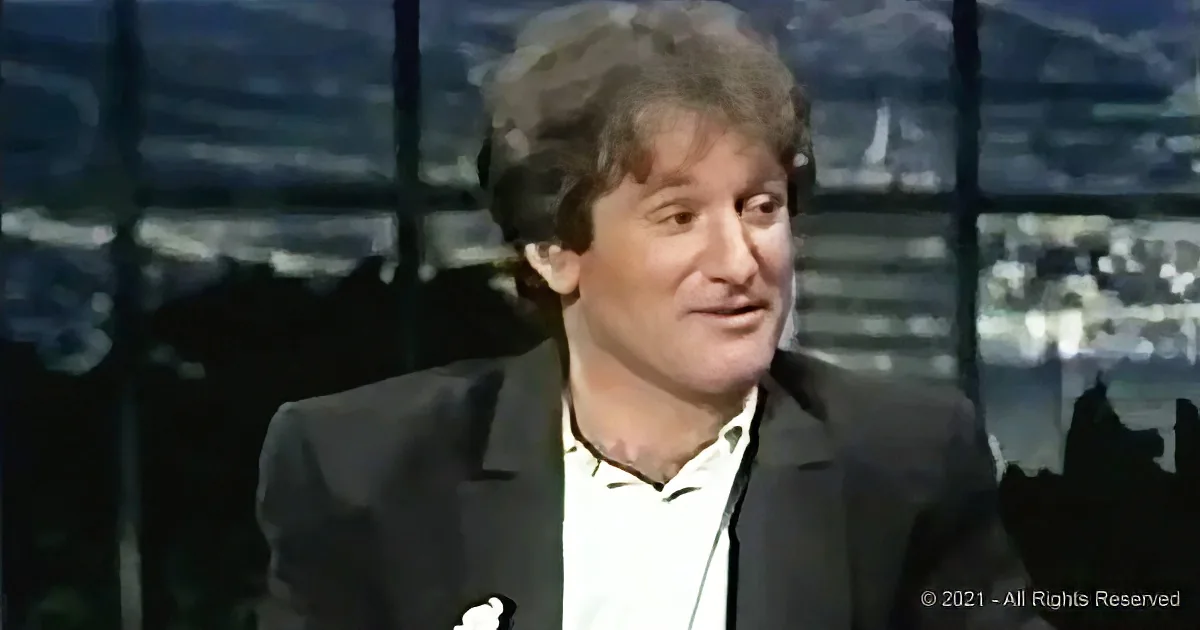 A man in a suit and white shirt is speaking, with a blurred background of a cityscape.