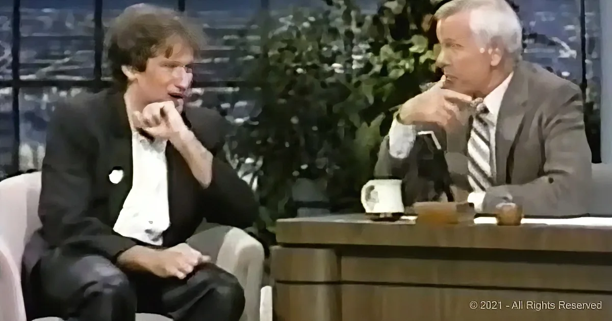 Two men in suits sit on a talk show set, one gesturing with his hand.