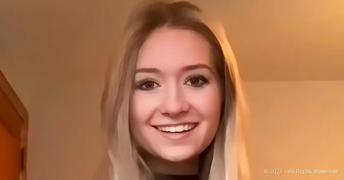 A smiling person with long hair, wearing a light-colored top, in a softly lit indoor setting.