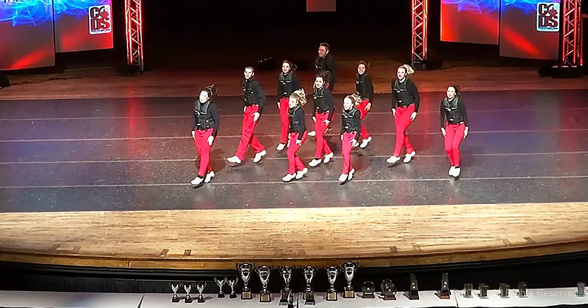 A group of dancers in red pants and black tops walk across a stage with trophies lined up below.