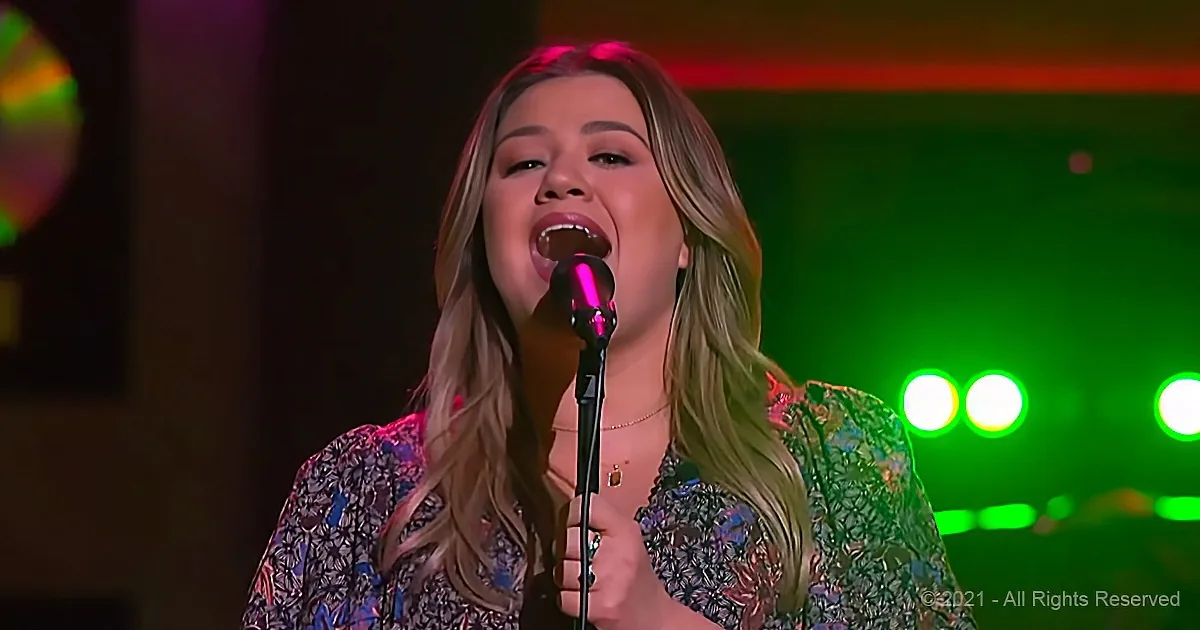 A woman sings into a microphone, wearing a floral dress, with green stage lights behind her.