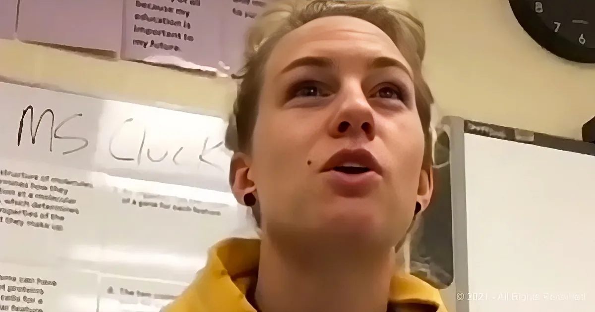 A person in a yellow hoodie looks up, with a whiteboard and clock in the background.