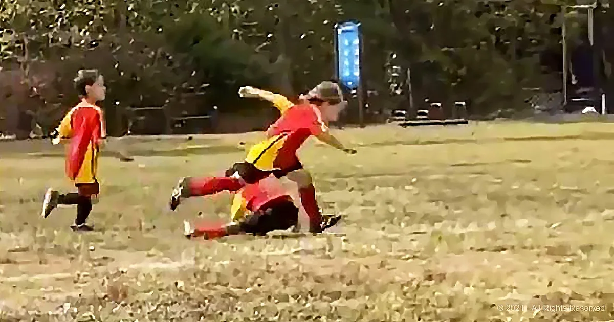 A child in red and yellow falls to the ground while another runs nearby on a grassy field.