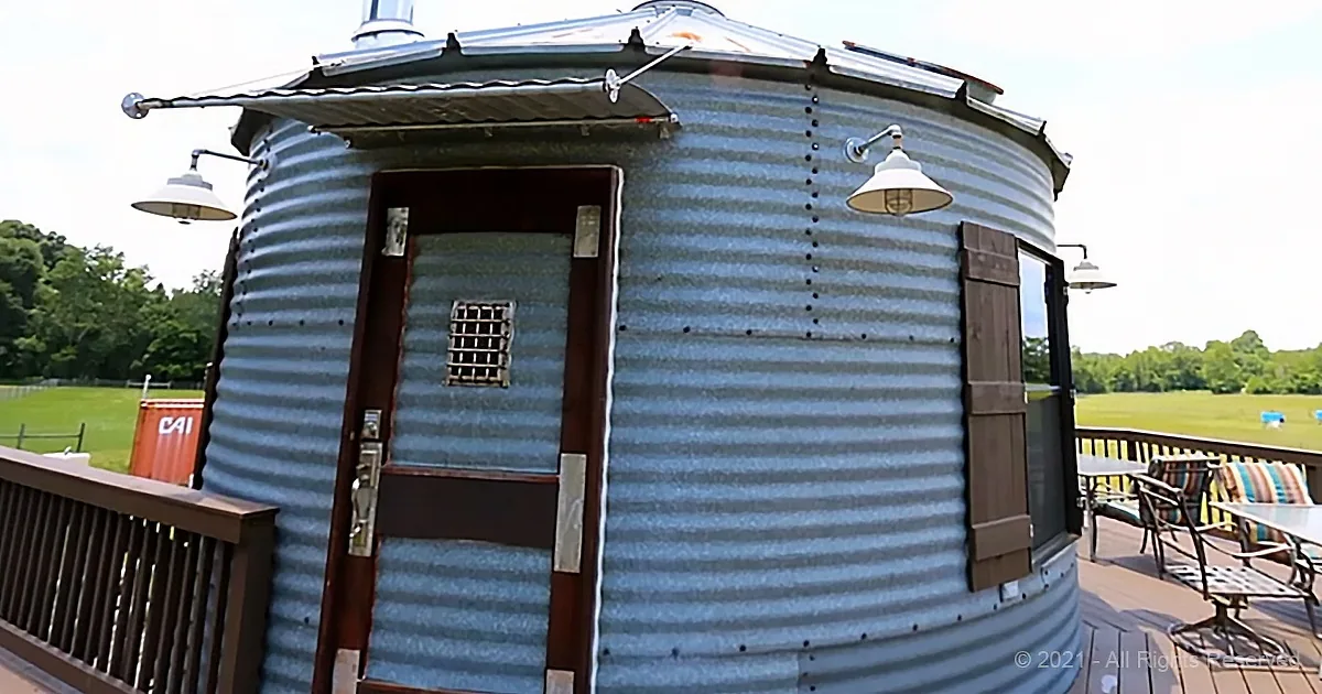 A corrugated metal silo-shaped structure with a door, windows, and outdoor lighting on a wooden deck.