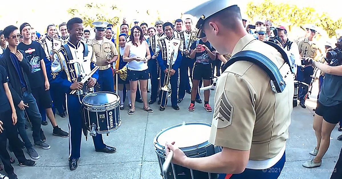 A marching band member in uniform plays a drum in front of a crowd, with other band members and spectators in the background.
