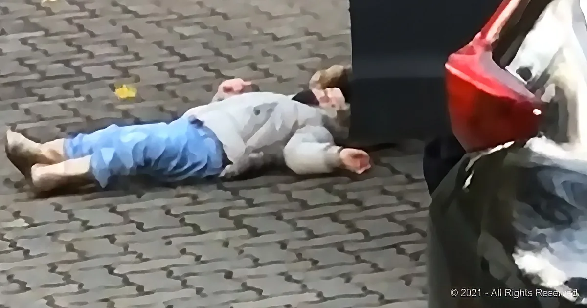A child lies on a cobblestone street near a parked car.