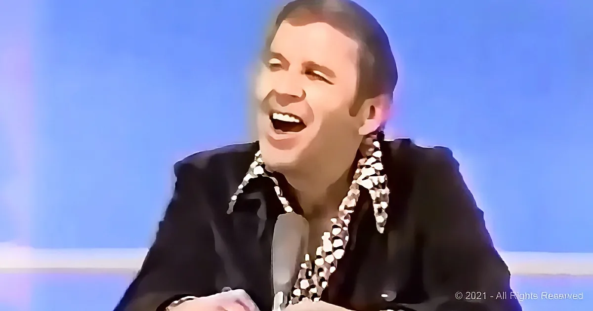 A man in a black suit and patterned shirt is smiling widely, with a bright blue background.