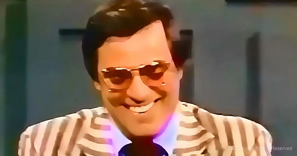 A man in a striped shirt smiles while wearing glasses, with a blurred background.