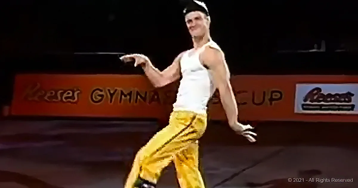A gymnast in a white tank top and yellow pants performs on a mat, with a Reese's Gymnastics Cup sign in the background.