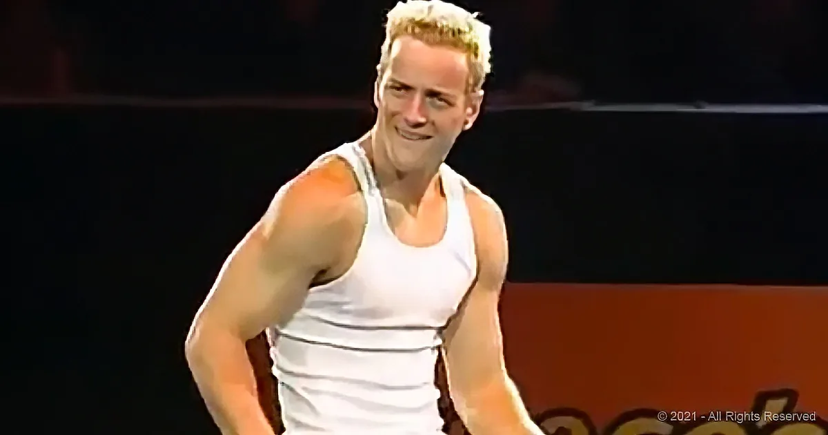 A man in a white tank top is smiling and looking to the side, with a dark background and some text in the corner.
