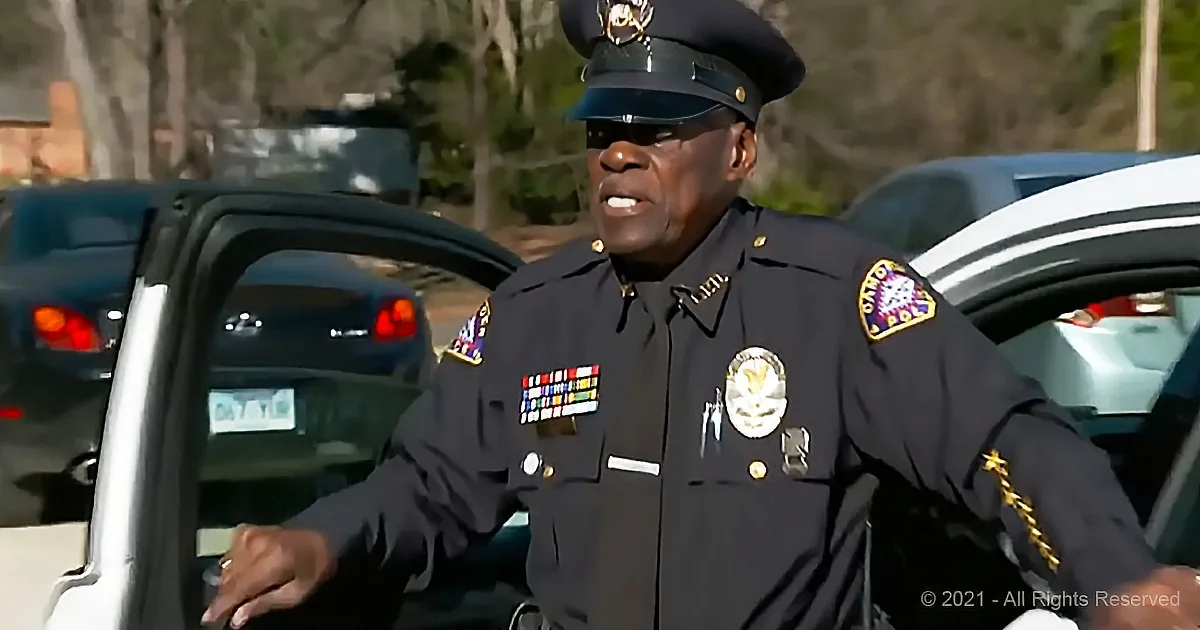 A police officer in uniform stands near a car, gesturing with his hands.