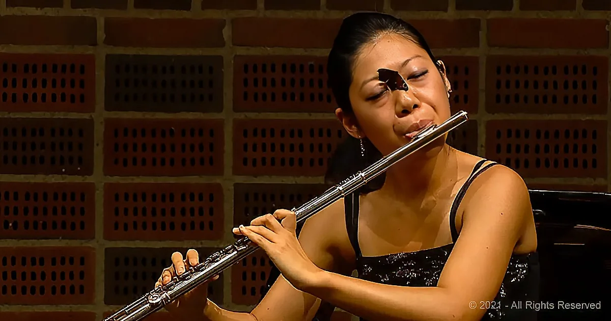 A woman in a black dress plays a flute against a brick wall backdrop.