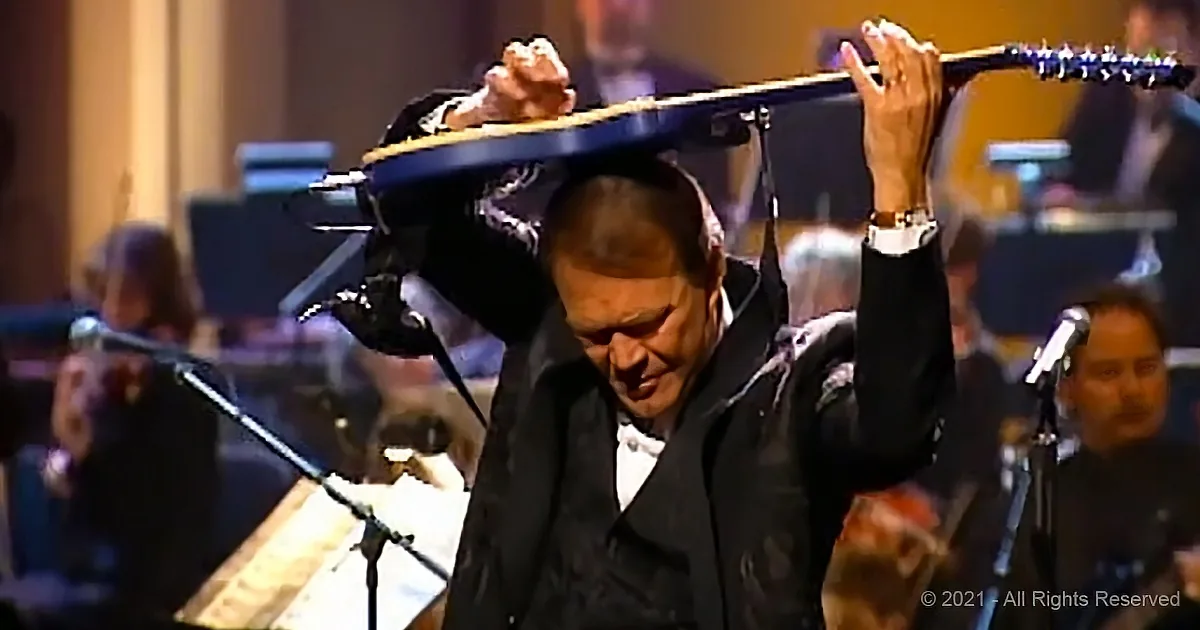 A man in a suit holds a guitar over his head, performing on stage with an orchestra in the background.