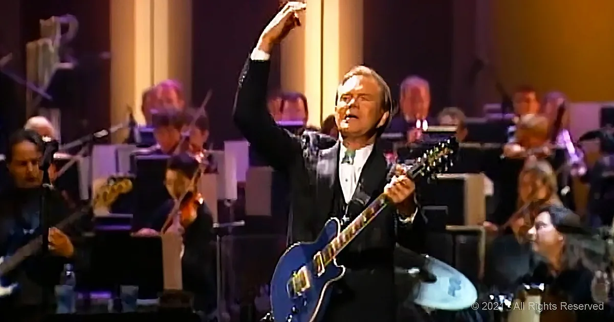 A man in a suit plays a blue guitar on stage with an orchestra in the background.