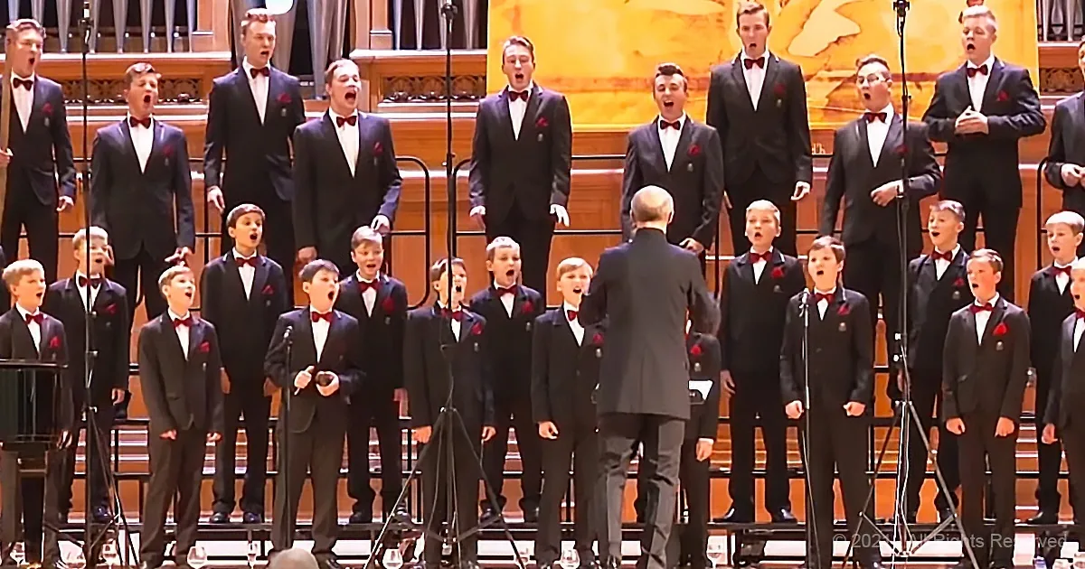A choir in formal attire sings under a conductor's direction in a grand hall.
