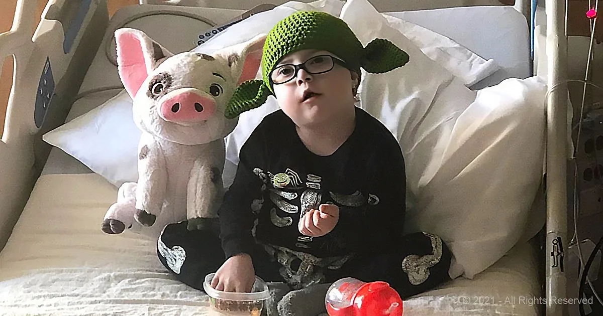 A child in a hospital bed wearing a green Yoda hat, surrounded by pillows and a stuffed pig.