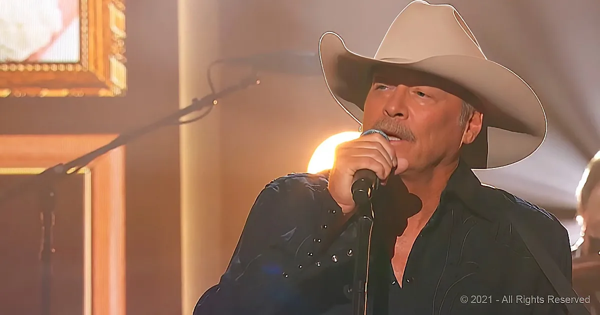 A man in a cowboy hat sings into a microphone, wearing a black shirt with studs, in a warmly lit setting.