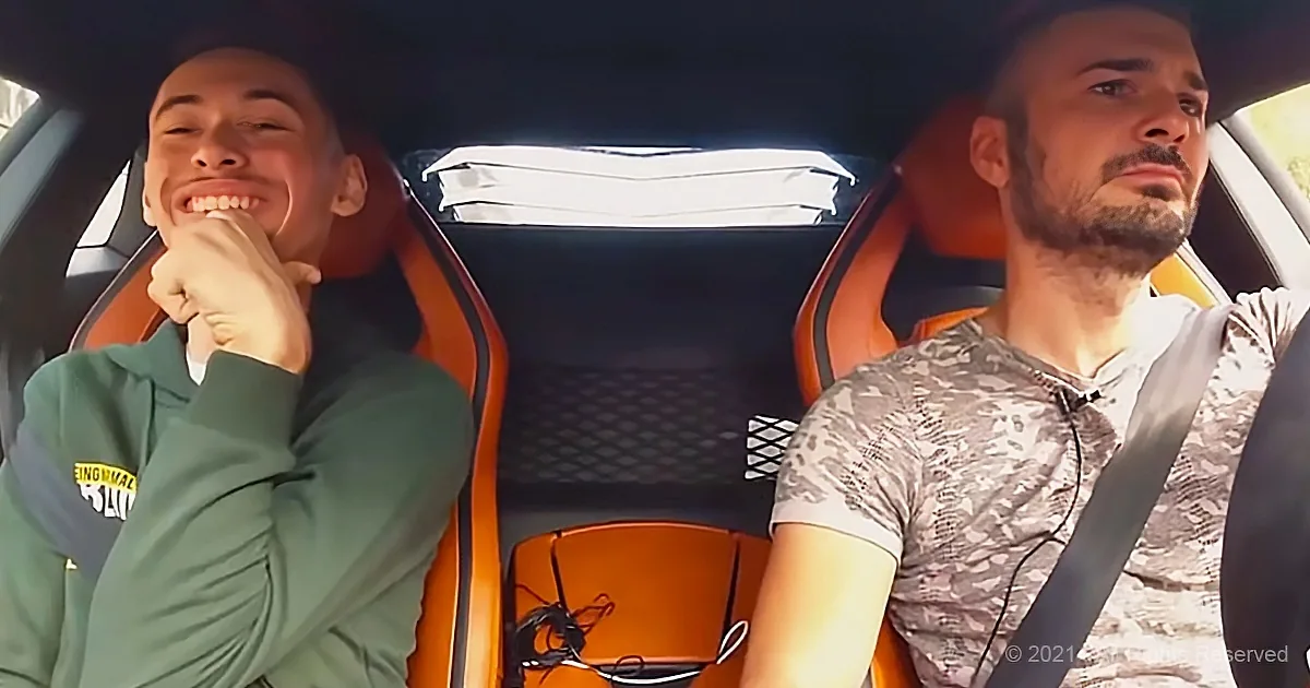 Two men are in a car, one smiling and wearing a green shirt, the other focused, wearing a camouflage shirt.