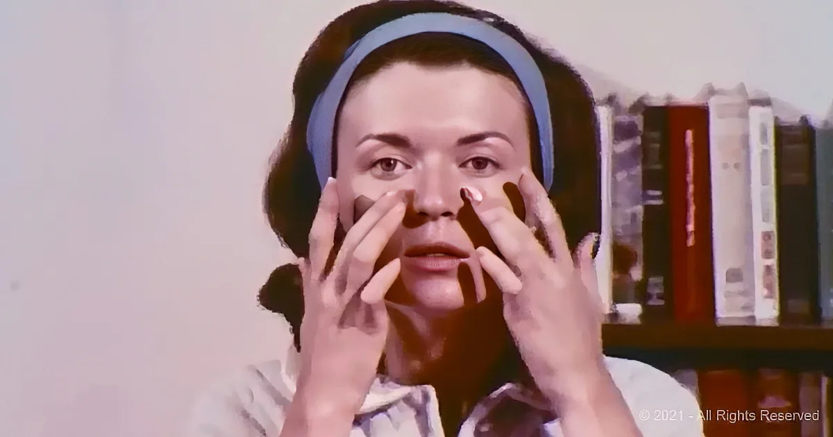 A woman wearing a blue headband and striped shirt is applying cream to her face with both hands.