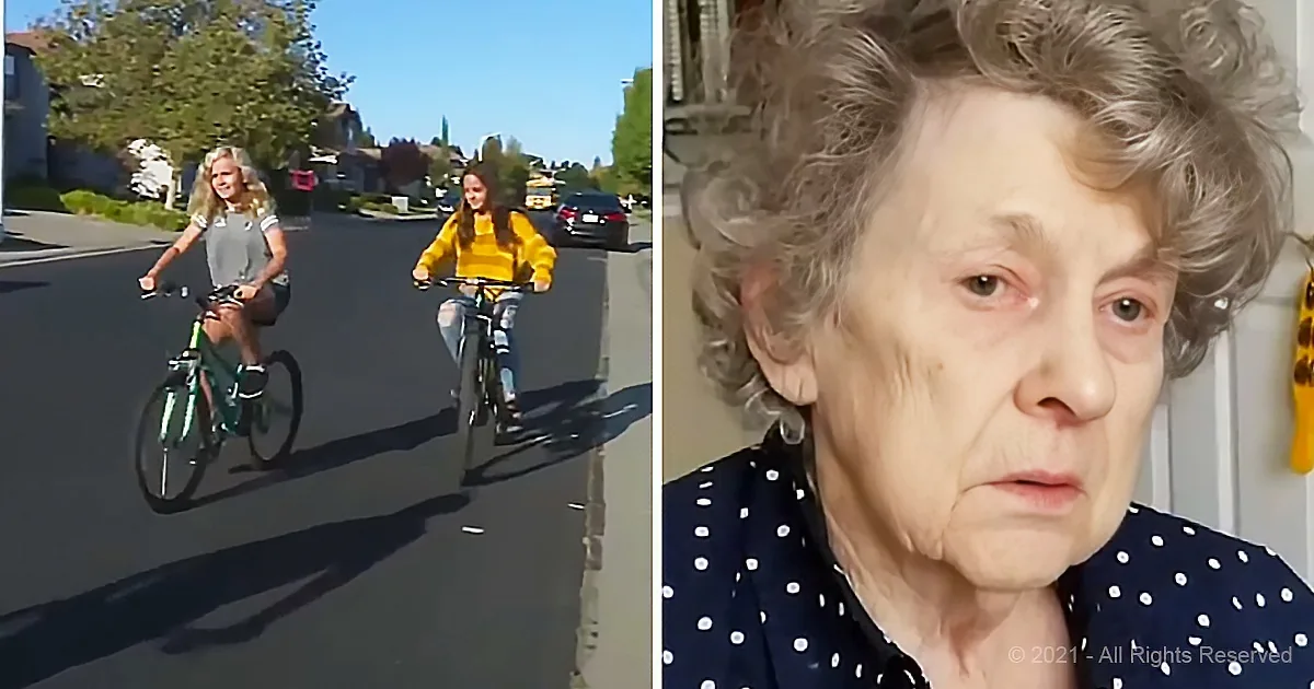 Two girls ride bicycles on a sunny street, while an elderly woman with gray hair looks on.