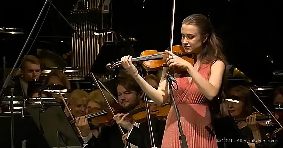 A violinist in a pink dress plays passionately in front of an orchestra.