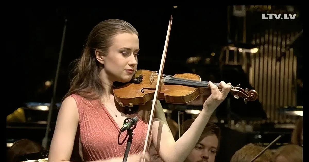 A woman in a red dress plays the violin on stage, surrounded by musicians and an audience.
