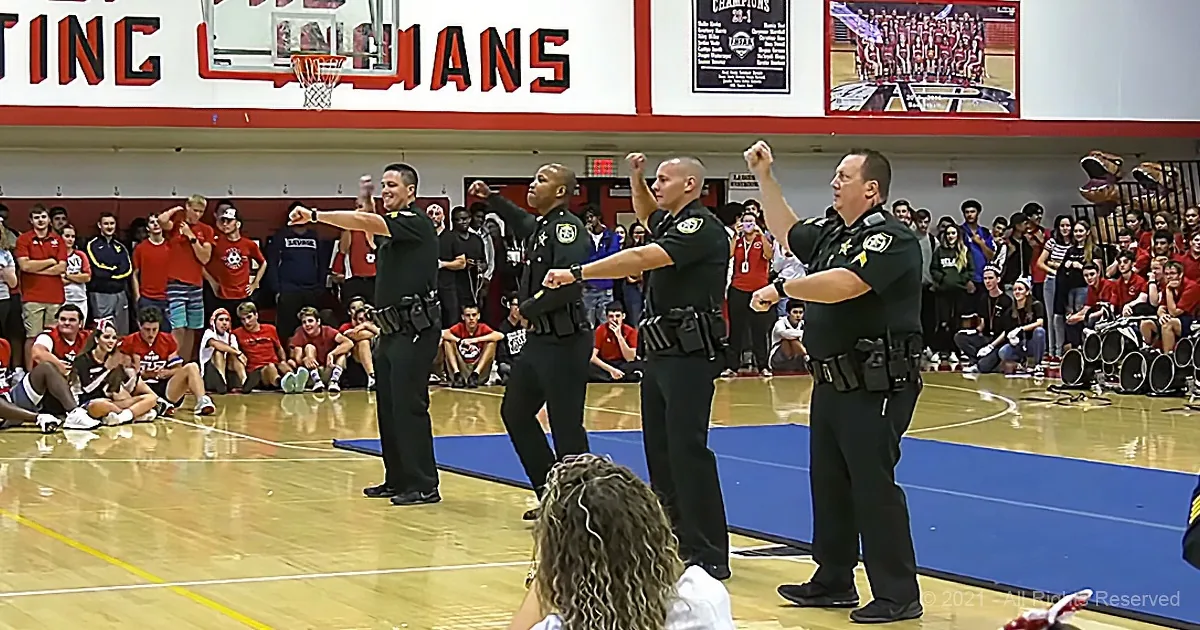 Four police officers in uniform perform a synchronized dance routine on a basketball court.